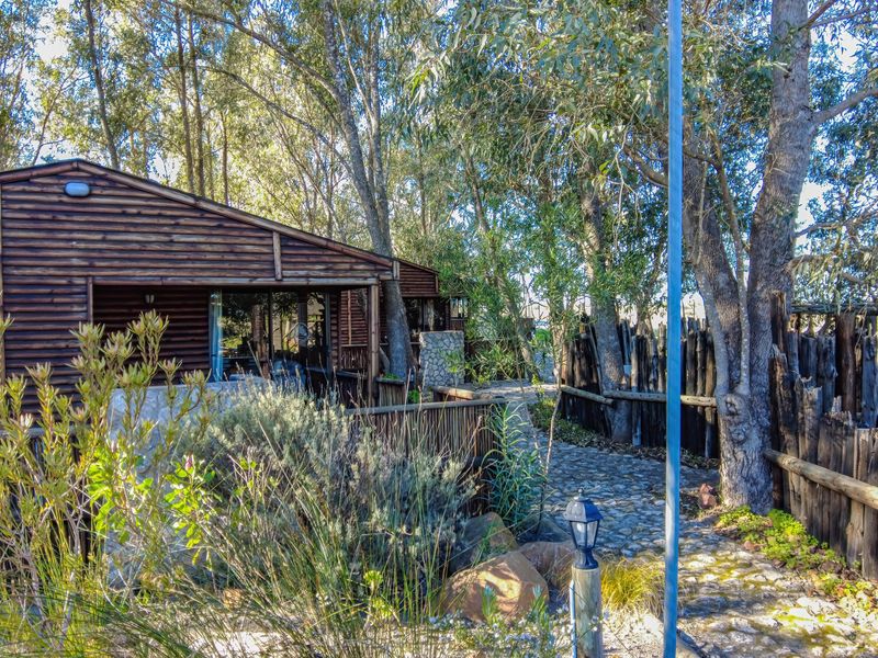 A rustic timber-clad farm dwelling featuring a stone pathway, natural landscaping, and traditional timber pole fencing in a wooded setting.