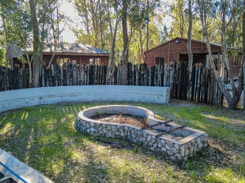 Outdoor entertainment area featuring a stone-clad fire pit, curved concrete seating, and rustic timber pole fencing set within a wooded farm garden.