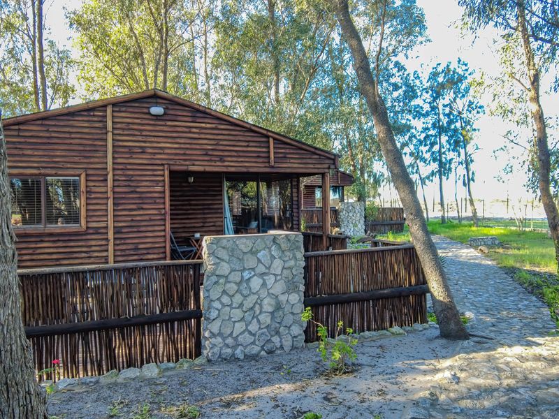 A rustic timber log cabin featuring a stone masonry pillar, timber pole fencing, and a stone paved pathway set within a wooded farm environment.
