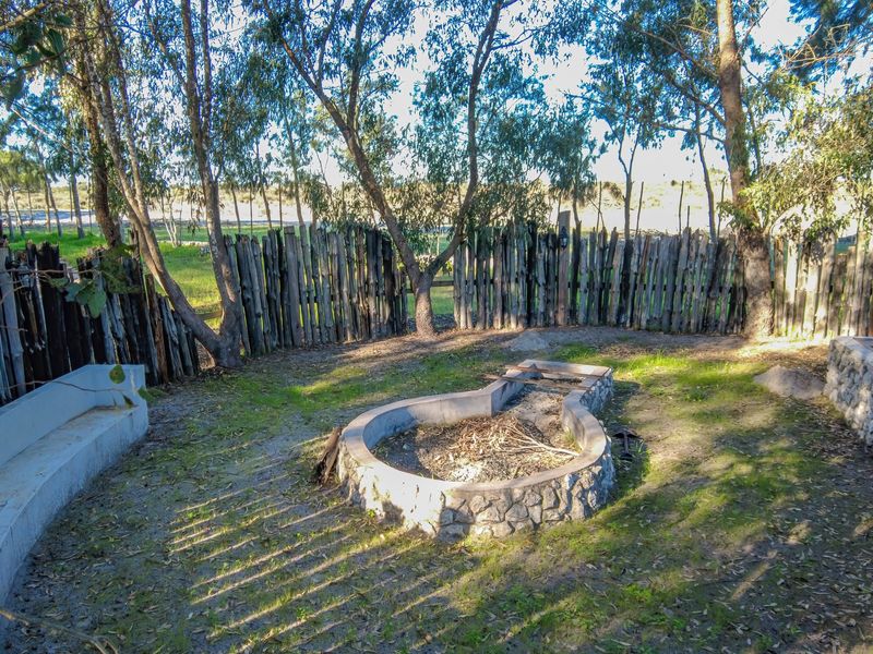 Outdoor garden area featuring a stone fire pit, built-in concrete bench seating, and rustic timber pole fencing set within a natural landscape.