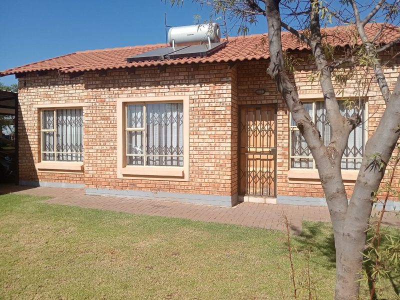 A well-maintained facebrick cluster home featuring a tiled roof, solar geyser, security gates, and a neat front garden with a paved walkway.