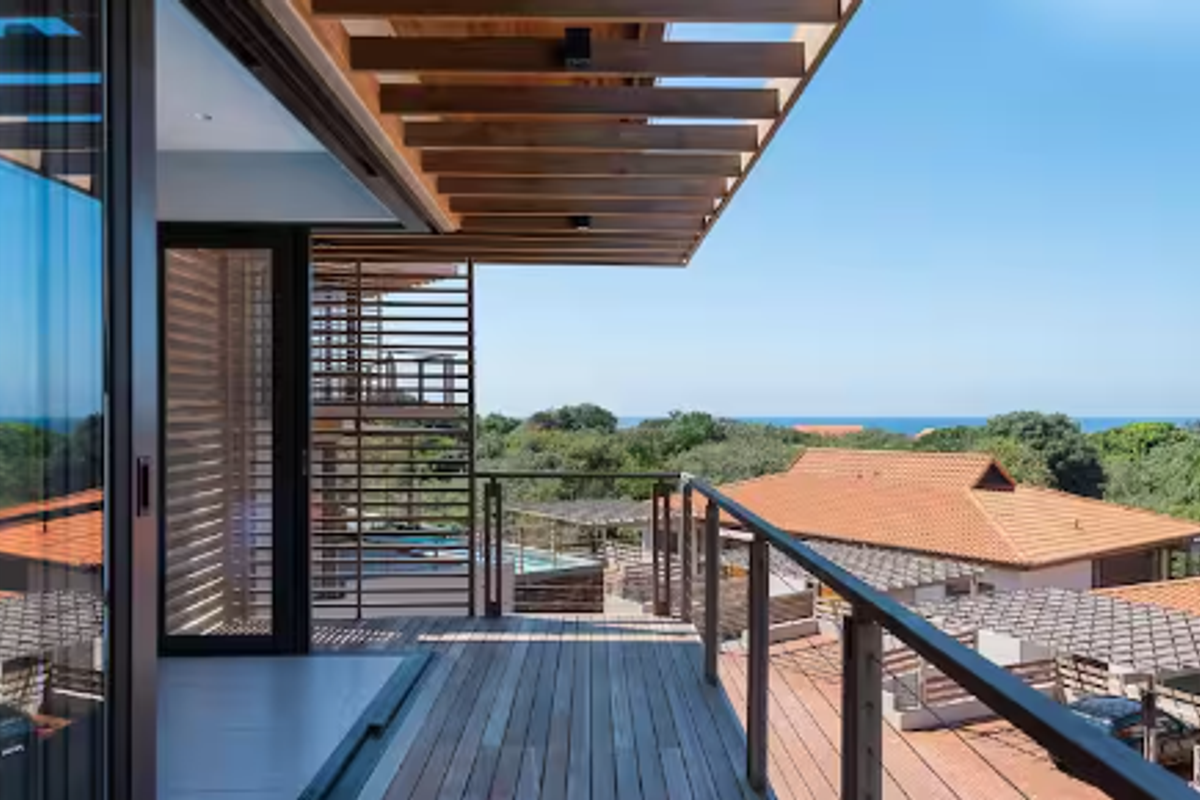 A modern, elevated timber balcony featuring a pergola roof, metal railings, and seamless access to the interior through large sliding glass doors.