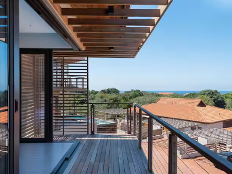 A modern, elevated timber balcony featuring a pergola roof, metal railings, and seamless access to the interior through large sliding glass doors.