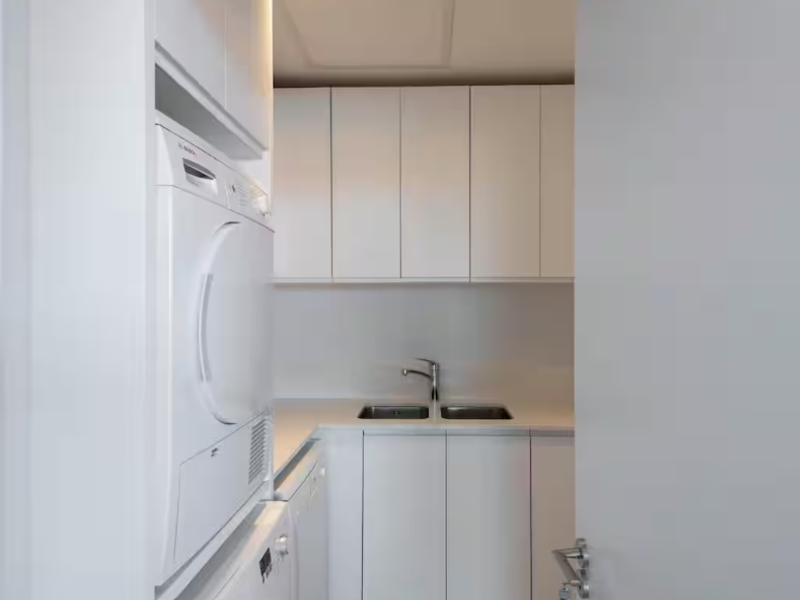 A clean, modern laundry room featuring white built-in cabinetry, a double sink, and stacked laundry appliances.