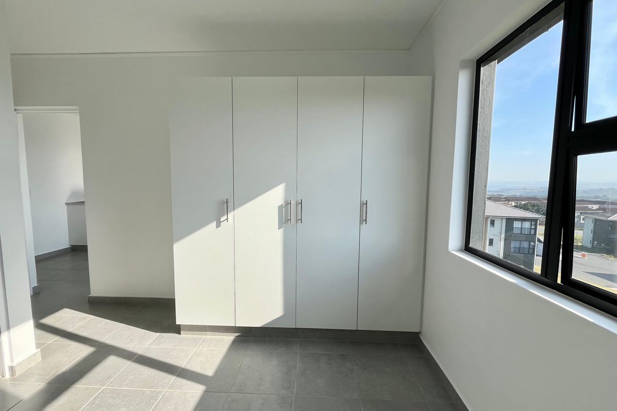 A bright, empty bedroom featuring modern built-in cupboards, neutral tiled flooring, and a large window with natural light and exterior views.