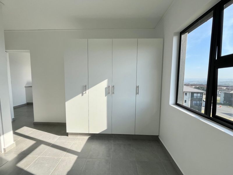 A bright, empty bedroom featuring modern built-in cupboards, neutral tiled flooring, and a large window with natural light and exterior views.
