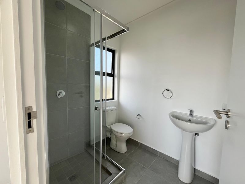 A clean, modern bathroom featuring a pedestal basin, toilet, and a glass-enclosed shower with grey floor and wall tiling.