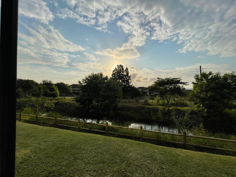 A scenic view of a natural stream and landscaped lawn, framed by a wooden post and rail fence under a sunset sky.
