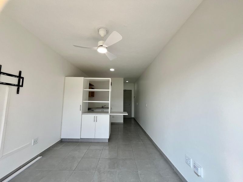 An open-plan living area featuring tiled flooring, a ceiling fan, and a compact kitchenette with built-in cabinetry.