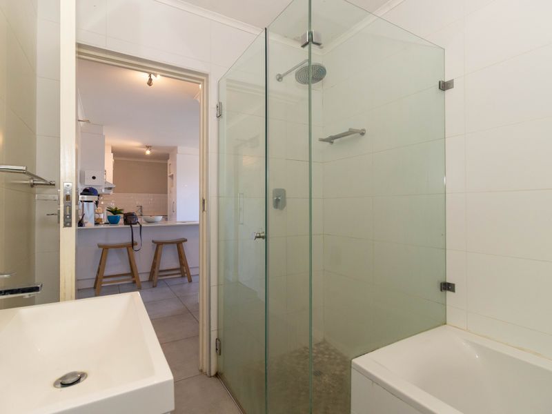 A clean, modern bathroom featuring a glass-enclosed shower, white basin, and neutral tiling, with a view through to the adjacent kitchen area.