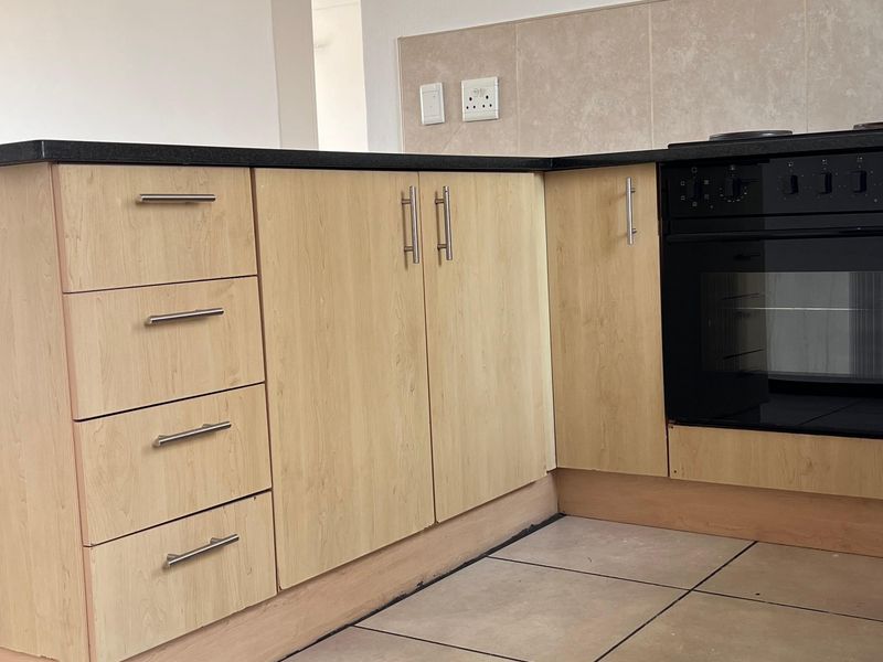 A functional kitchen area featuring light wood-finish cabinetry, a built-in oven, tiled splashback, and neutral floor tiles in an open-plan layout.