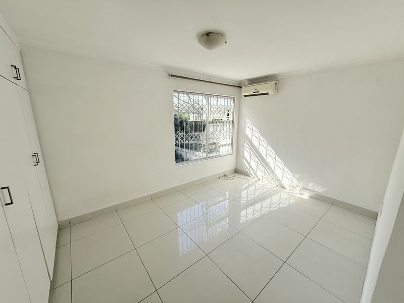 A bright, empty bedroom featuring white tiled flooring, built-in cupboards, and a wall-mounted air conditioning unit with security bars on the window.