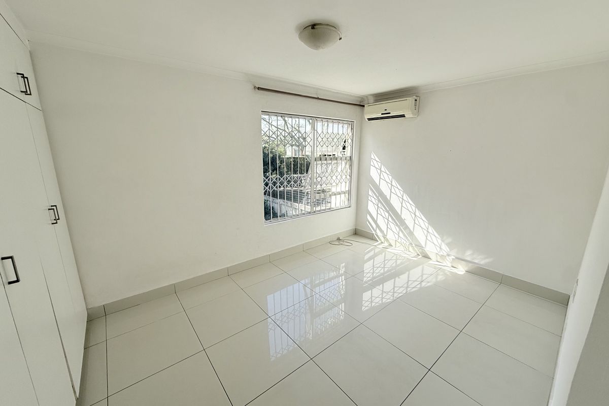 A bright, empty bedroom featuring white tiled flooring, built-in cupboards, and a wall-mounted air conditioning unit with security bars on the window.