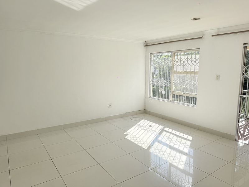 A bright, empty living area featuring white walls, tiled flooring, and security bars on the windows and door.