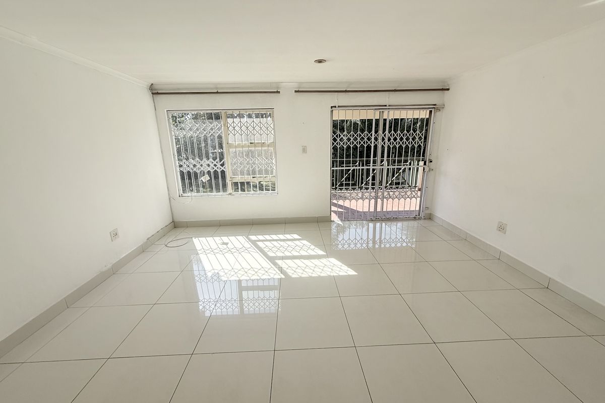 A bright, empty living room featuring white tiled flooring, large windows with security bars, and a sliding door leading to an outdoor area.