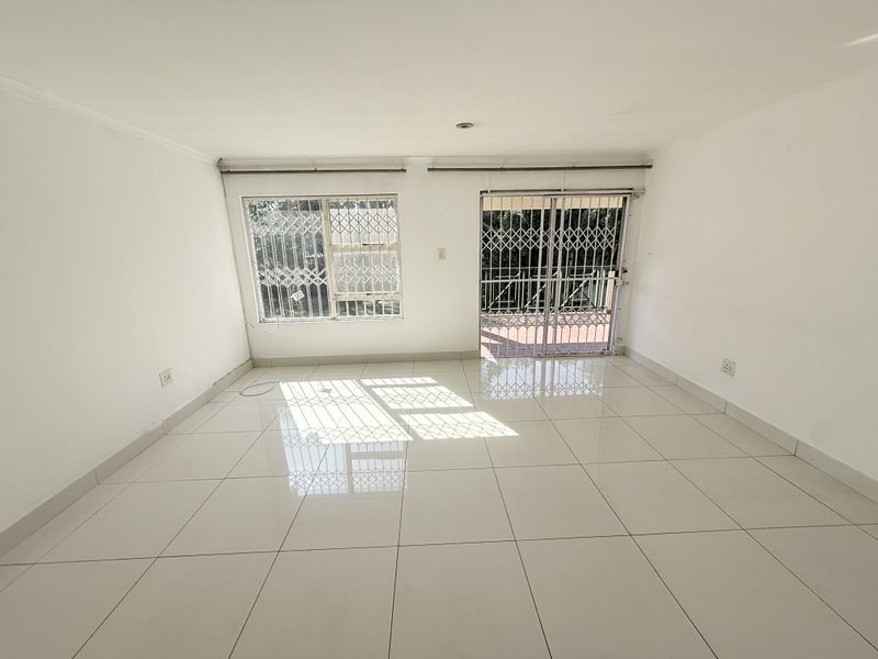 A bright, empty living room featuring white tiled flooring, large windows with security bars, and a sliding door leading to an outdoor area.