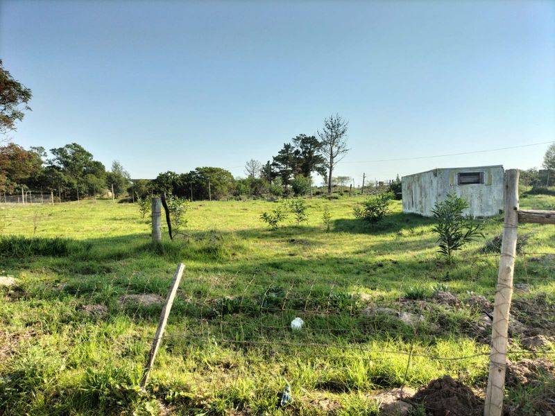 A vacant residential plot featuring grassy terrain, perimeter wire fencing with wooden posts, and a small weathered utility structure.