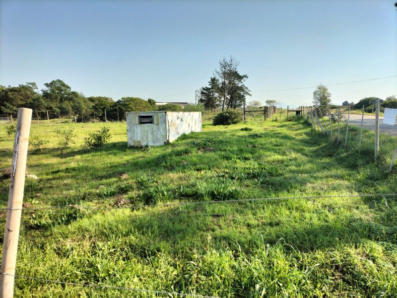 A view of a vacant residential plot featuring wire fencing, overgrown grass, and a small, weathered storage structure on the property.
