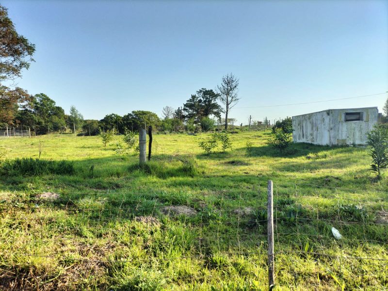 A vacant residential plot featuring grassy terrain, wire perimeter fencing, and a small concrete outbuilding structure.