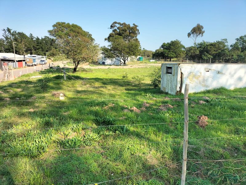 A view of a vacant residential plot featuring grassy terrain, perimeter wire fencing, and a small, weathered storage structure.