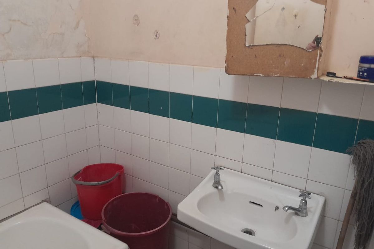 A dated bathroom featuring a wall-mounted basin with dual-handle taps and tiled splashbacks, requiring significant cosmetic renovation and repair to wall finishes.
