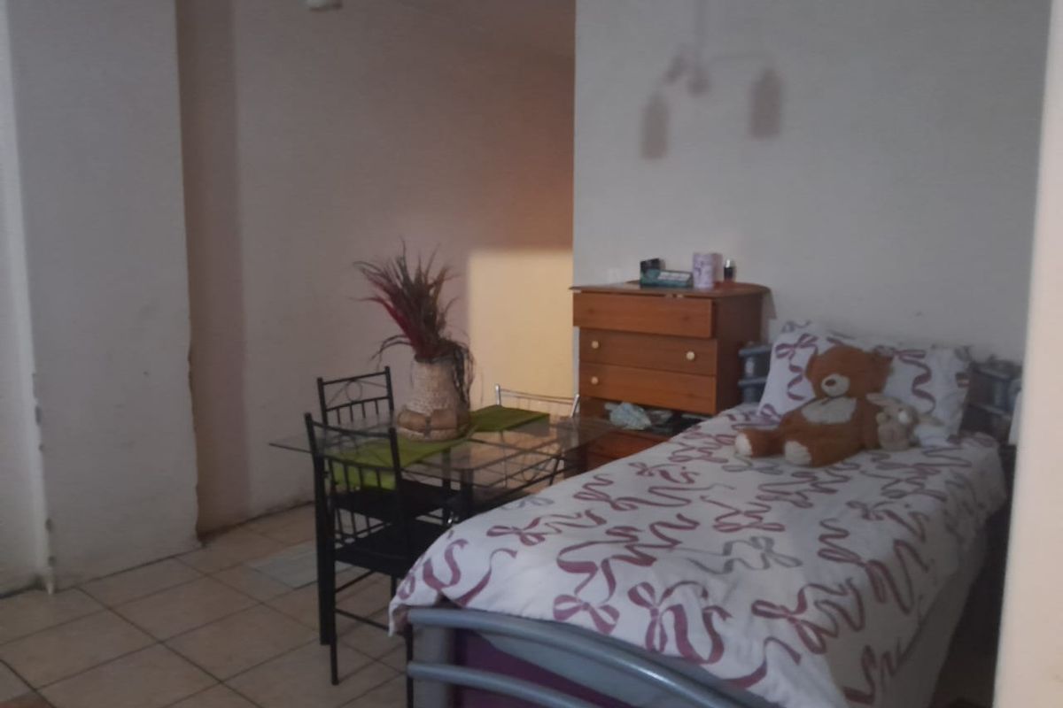 A bedroom featuring tiled flooring and plain walls, currently furnished with a single bed and small dining set.