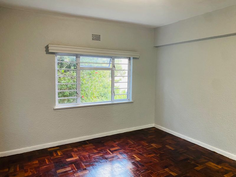 A bright, empty bedroom featuring polished parquet flooring, neutral walls, and a window fitted with security bars overlooking greenery.
