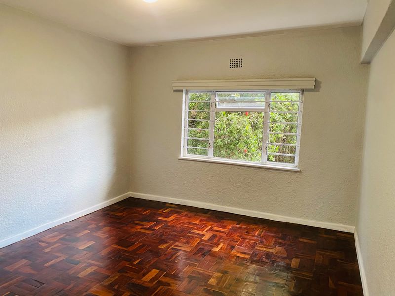 A bright, empty bedroom featuring classic parquet flooring, neutral walls, and a window fitted with security bars overlooking greenery.