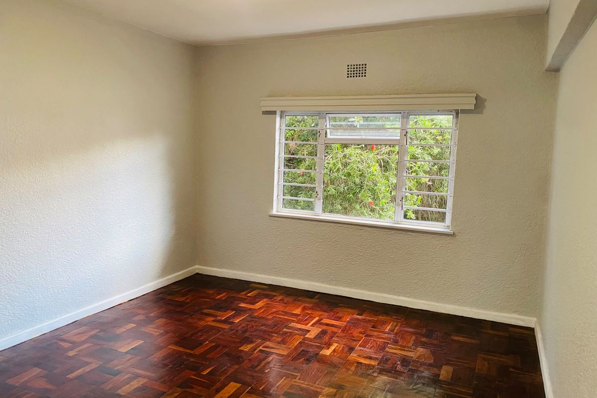 A bright, empty bedroom featuring classic parquet flooring, neutral walls, and a window fitted with security bars overlooking greenery.