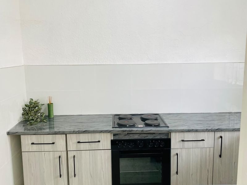 A clean, modern kitchen area featuring light wood-grain cabinetry, a dark stone-look countertop, and a built-in electric oven and hob with a tiled splashback.