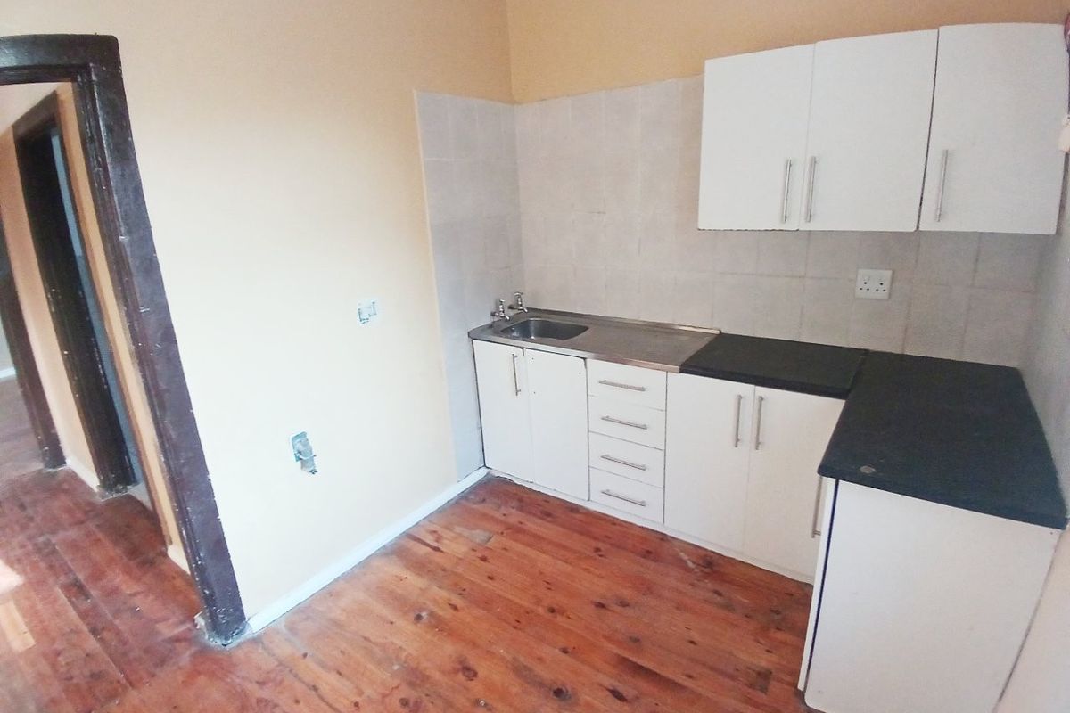 A compact kitchen featuring white cabinetry, dark laminate countertops, a stainless steel sink, and original timber flooring, ready for modernization.