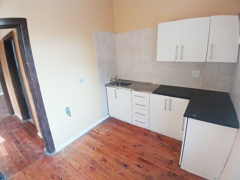 A compact kitchen featuring white cabinetry, dark laminate countertops, a stainless steel sink, and original timber flooring, ready for modernization.