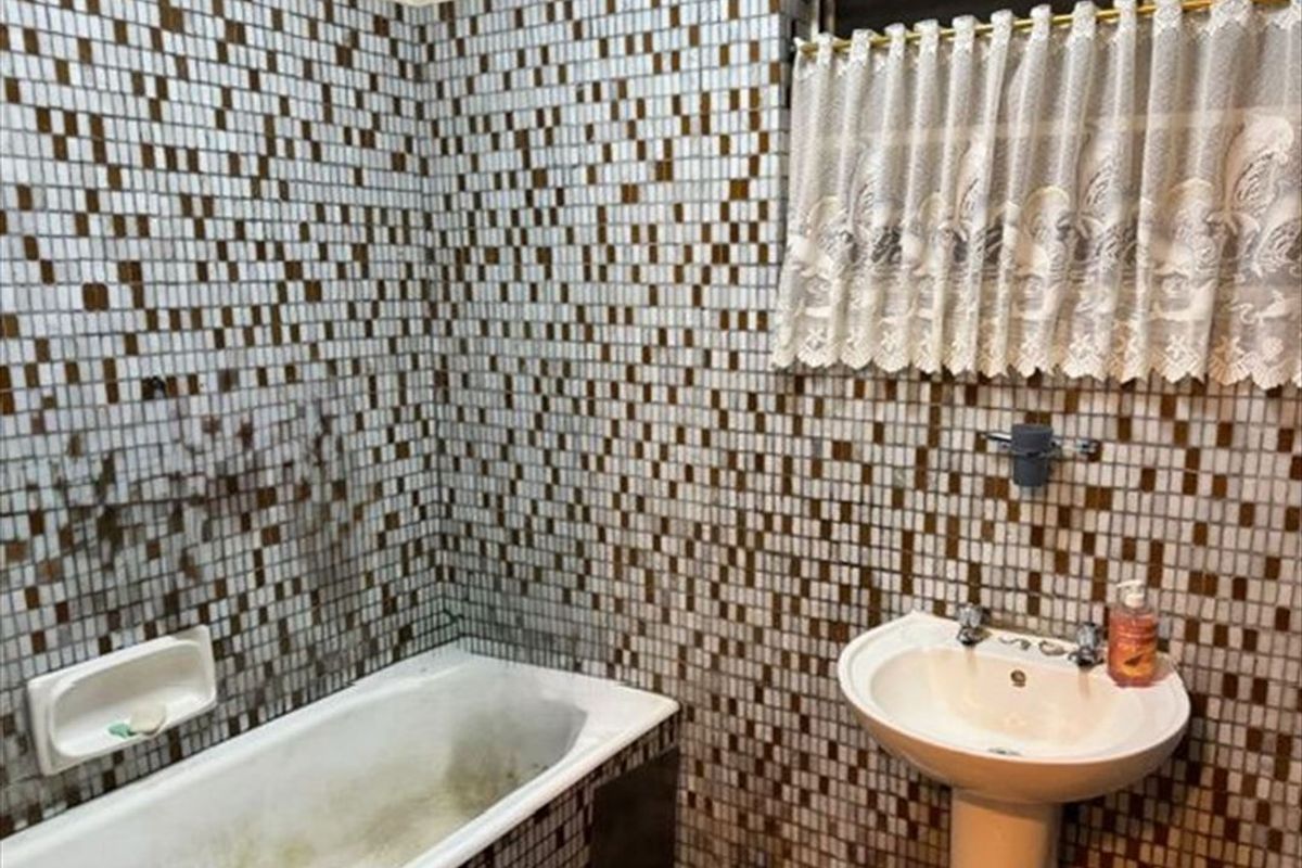 A dated bathroom featuring a pedestal basin and built-in bathtub with extensive mosaic wall tiling, requiring significant cleaning and refurbishment of fixed finishes.