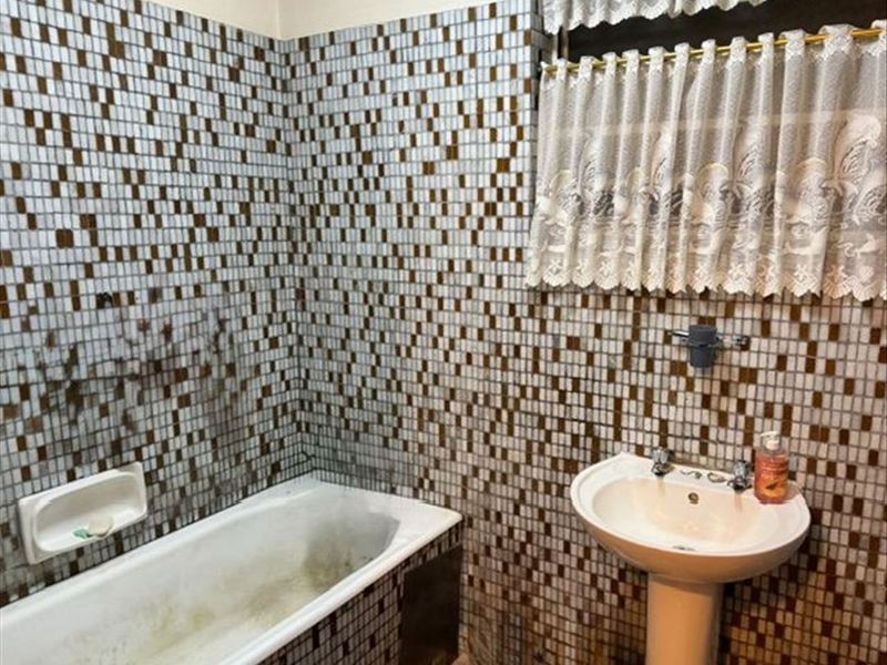 A dated bathroom featuring a pedestal basin and built-in bathtub with extensive mosaic wall tiling, requiring significant cleaning and refurbishment of fixed finishes.