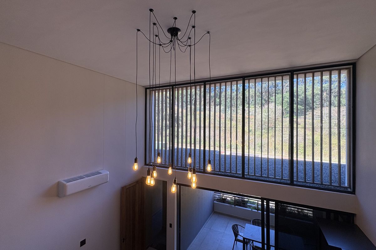 Spacious double-volume living area featuring large windows with vertical louvers, a modern multi-pendant light fixture, and a wall-mounted split-system air conditioner.