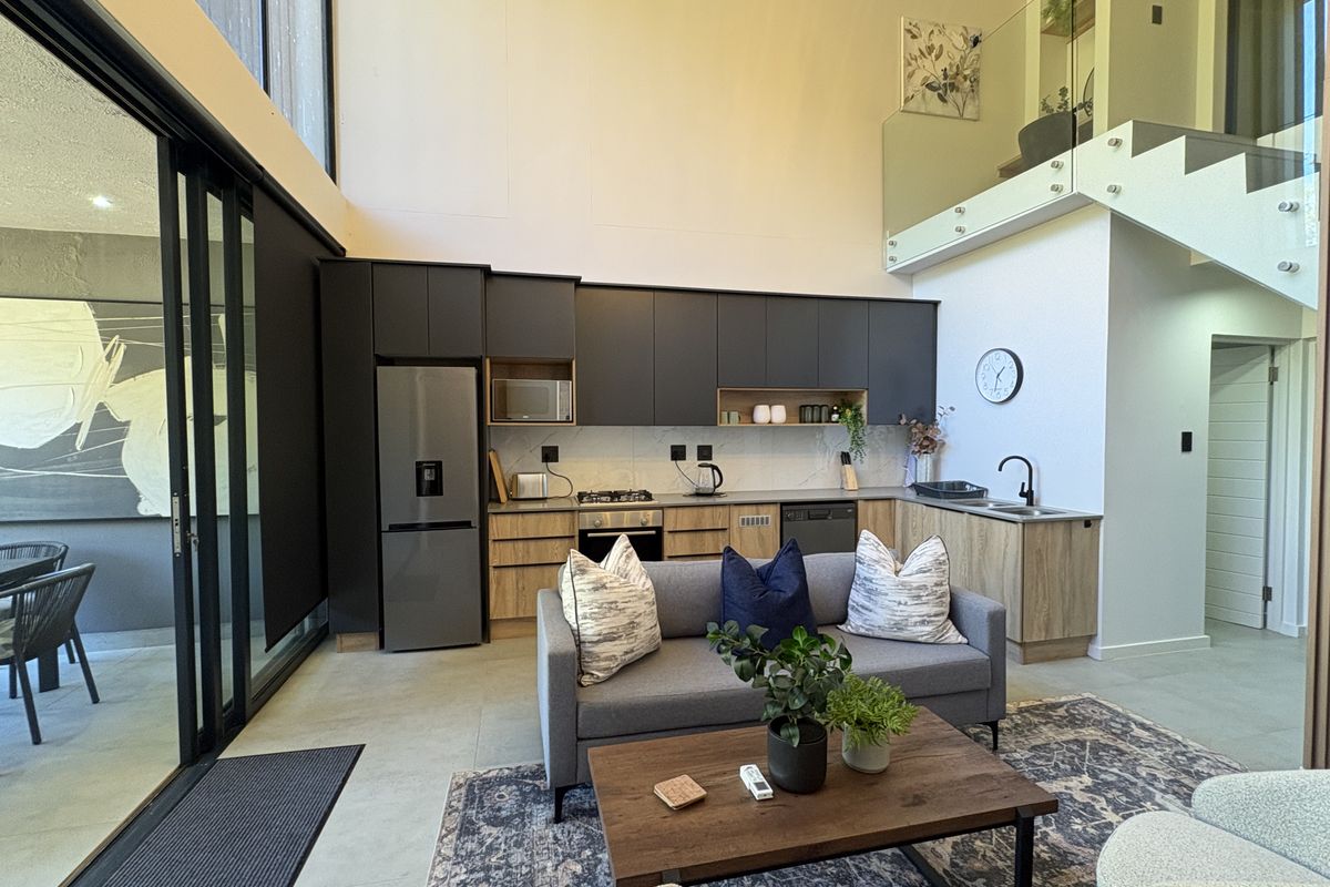 Modern open-plan living area featuring a sleek kitchen with dark cabinetry, integrated appliances, high ceilings, and a glass-railed staircase leading to a mezzanine level.