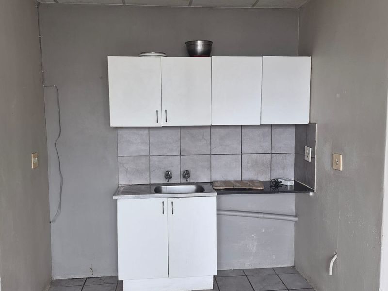 A compact, basic kitchen area featuring white cabinetry, a stainless steel sink, tiled backsplash, and tiled flooring, currently in need of cosmetic updates.
