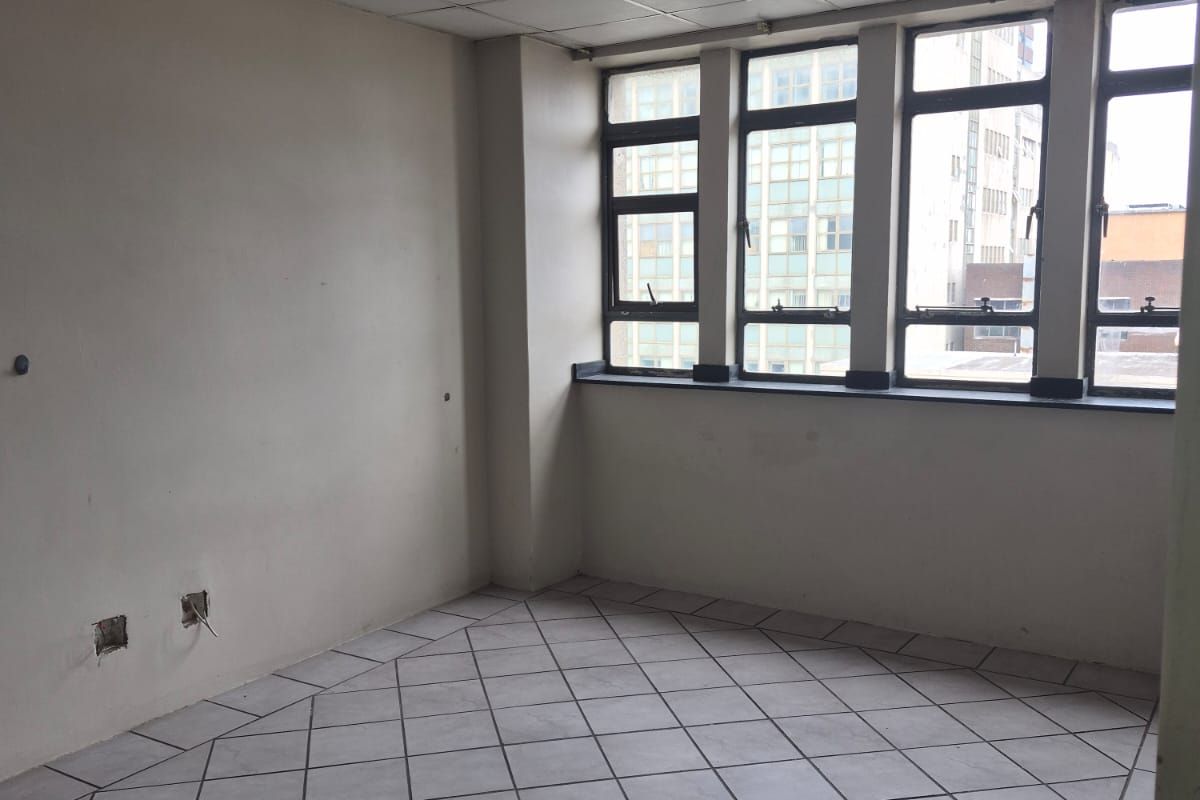 An empty living area featuring tiled floors, a suspended ceiling, and large windows providing natural light. The space requires minor cosmetic updates.