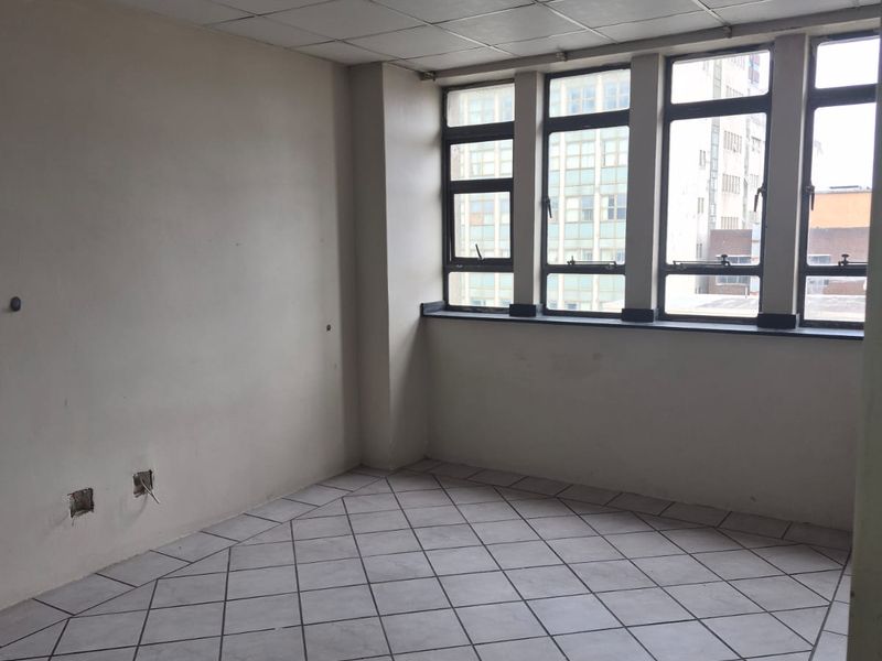An empty living area featuring tiled floors, a suspended ceiling, and large windows providing natural light. The space requires minor cosmetic updates.