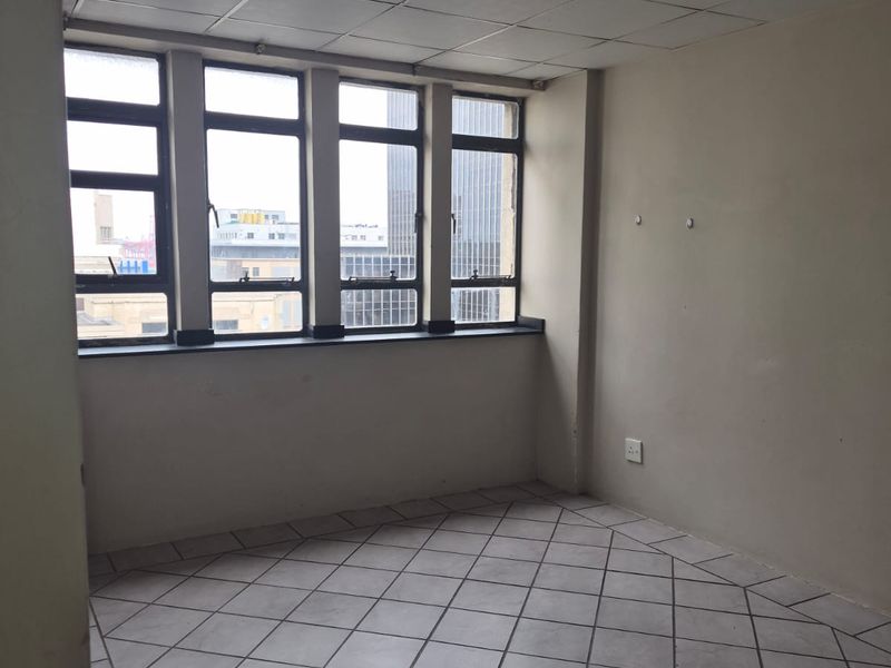 An empty living area featuring tiled flooring, a drop ceiling, and large windows providing natural light and urban views.