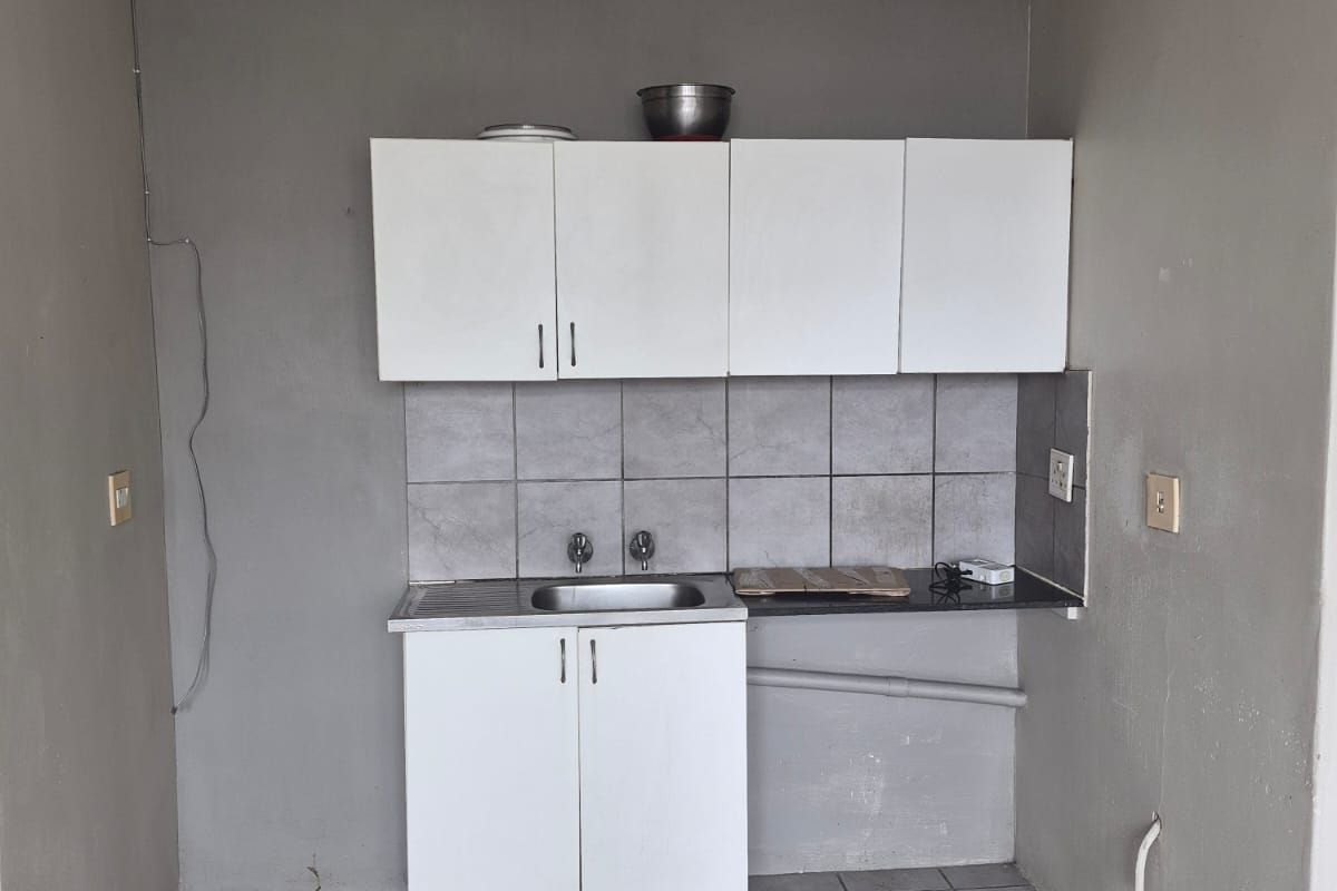 A compact, basic kitchen area featuring white cabinetry, a stainless steel sink, tiled backsplash, and tiled flooring, currently in need of cosmetic updates.