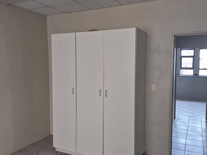 A sparse bedroom featuring tiled flooring, a drop ceiling, and a built-in white cupboard, with an adjacent room visible through the doorway.