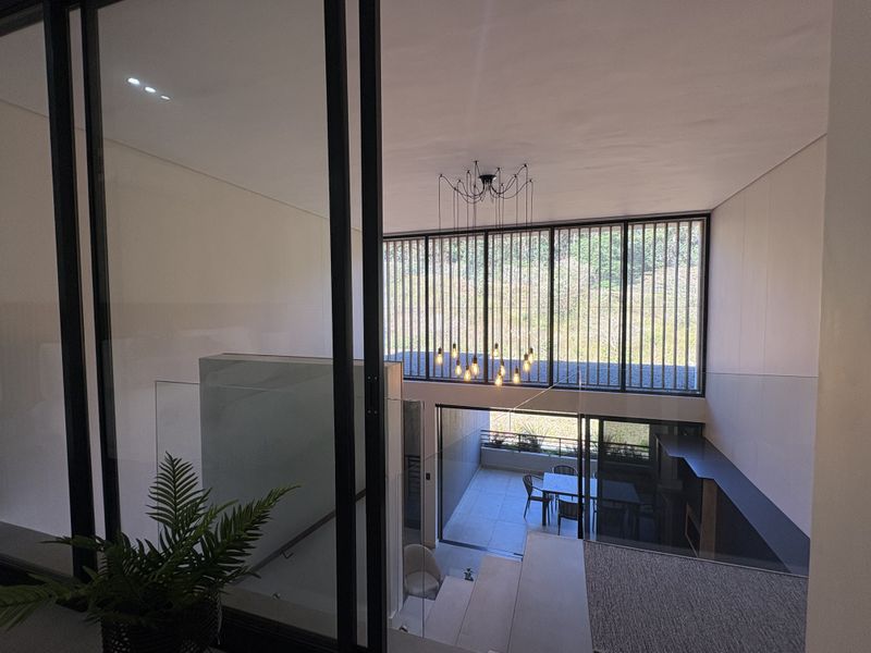 Modern open-plan living area featuring high ceilings, a striking multi-pendant chandelier, glass balustrades, and large windows overlooking a private balcony.
