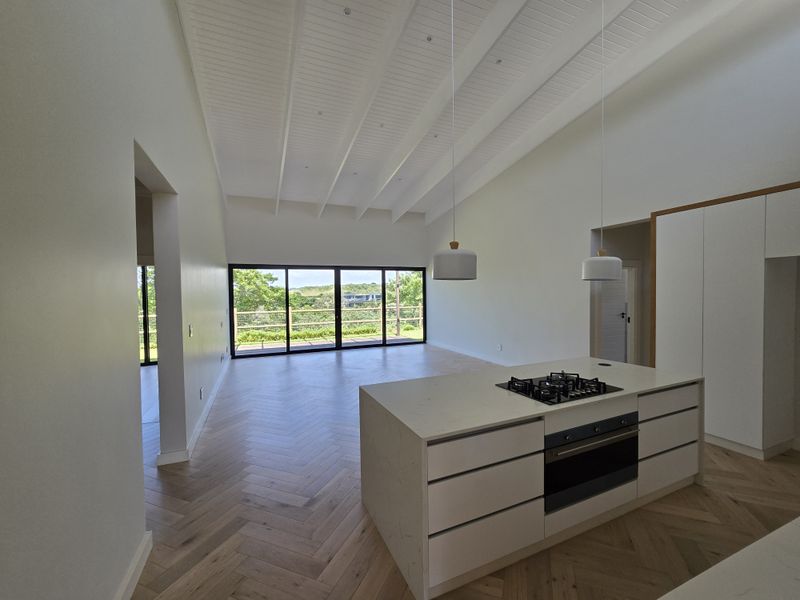 Spacious open-plan living area featuring high vaulted ceilings, herringbone timber flooring, and a modern kitchen island with integrated appliances, opening to a scenic outdoor view.