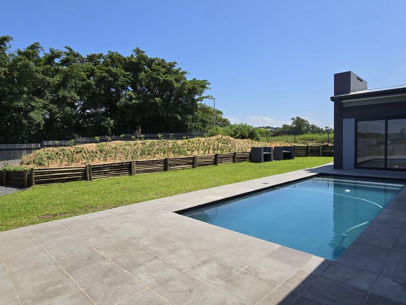 A well-maintained outdoor pool area featuring a tiled deck, built-in braai, and a landscaped garden with a retaining wall.