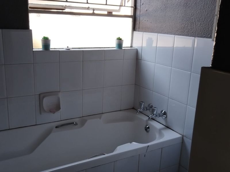 A functional bathroom featuring a built-in bathtub with chrome fixtures, white wall tiling, and a window with security bars.