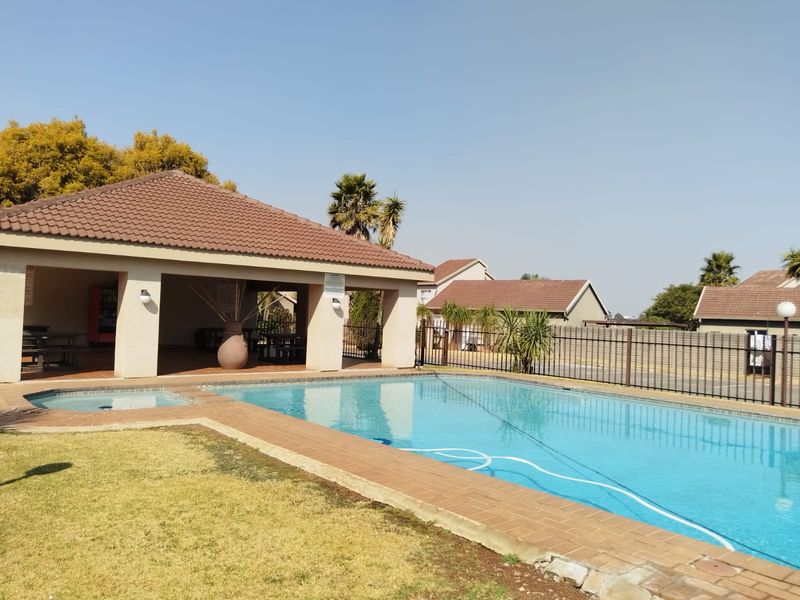 Communal pool area featuring a large swimming pool, paved surround, and a covered patio structure within a fenced complex.