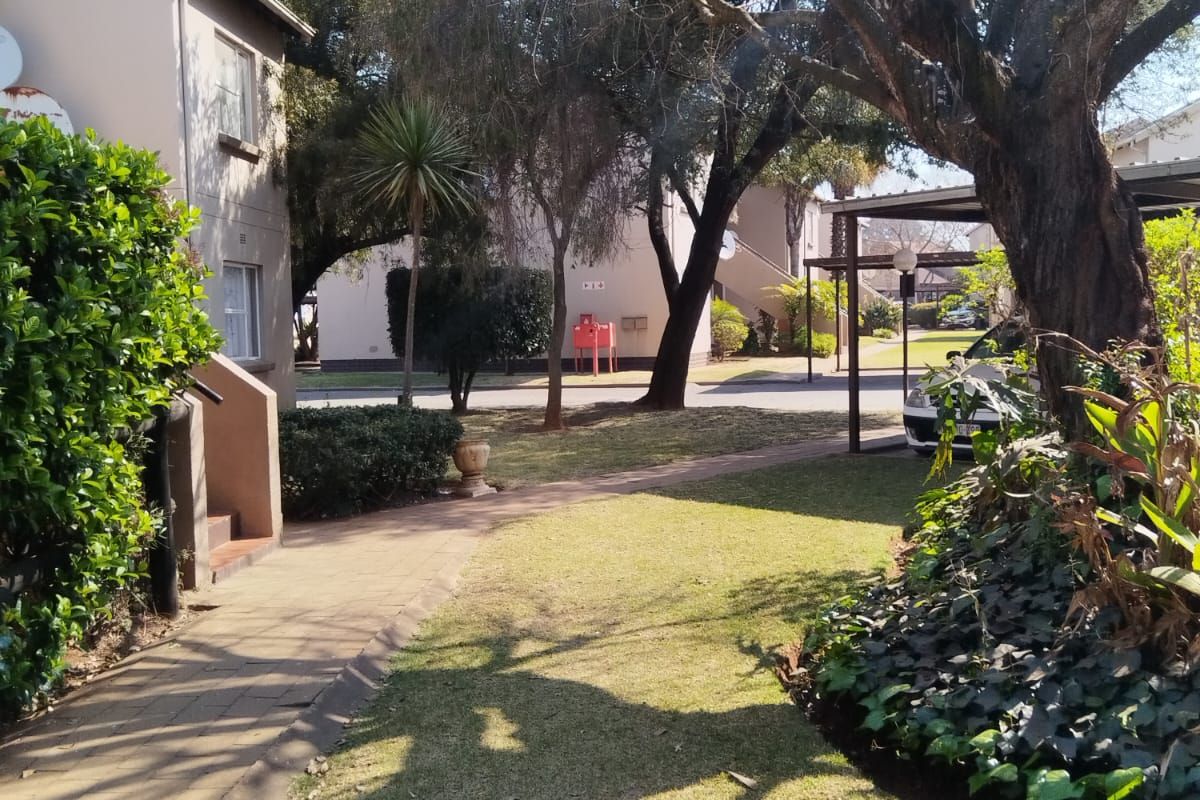 A view of the apartment complex exterior featuring a paved walkway, well-maintained garden area, and a covered carport structure.