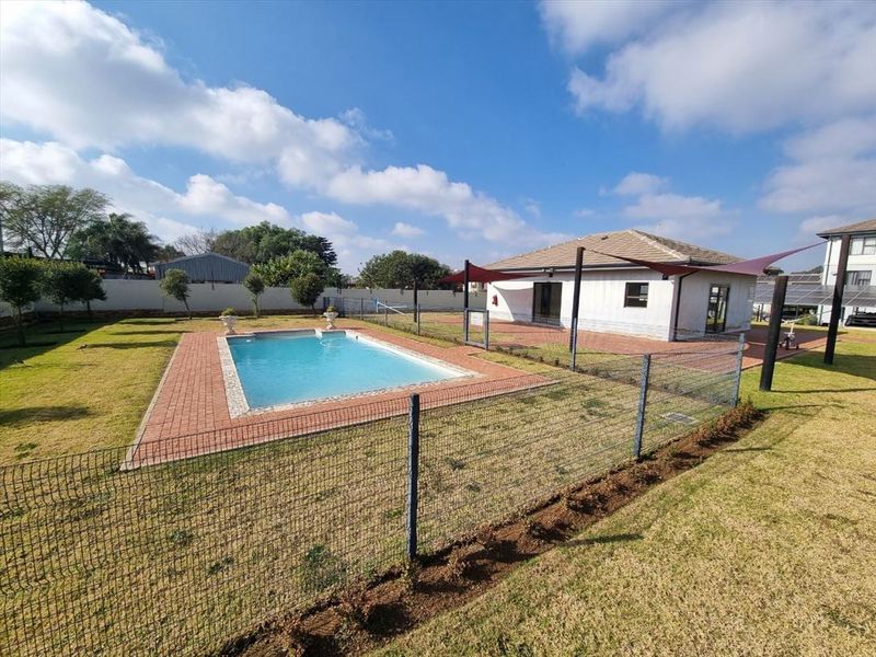 Outdoor pool area featuring a swimming pool with brick surround, shade sail, and perimeter fencing set within a landscaped garden.