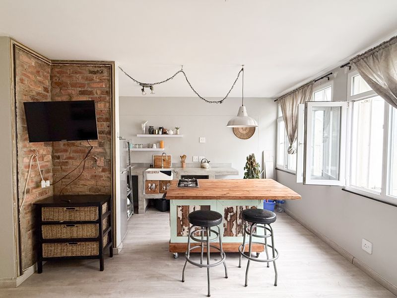 Open-plan living and kitchen area featuring an exposed brick wall, central kitchen island with cooktop, and large windows providing ample natural light.